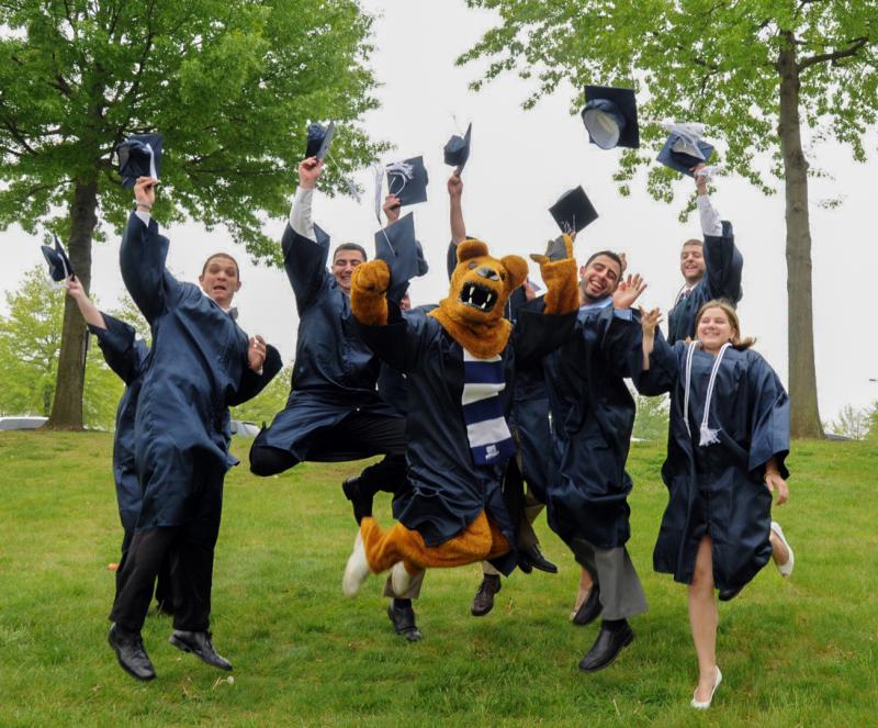 A group of graduating students in caps & gowns jump in the air in front of the Nittany Lion shrine