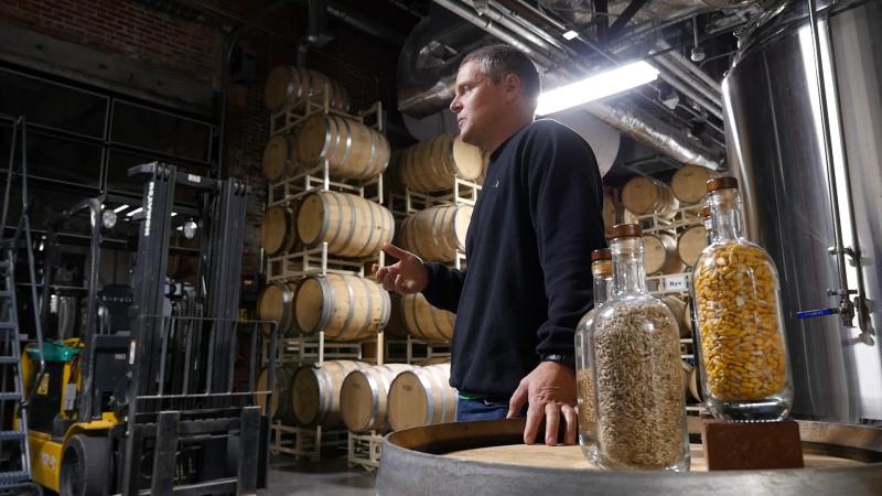 Kevin Lloyd stands in the Big Spring Distillery with grains in the foreground and aging barrels in the background